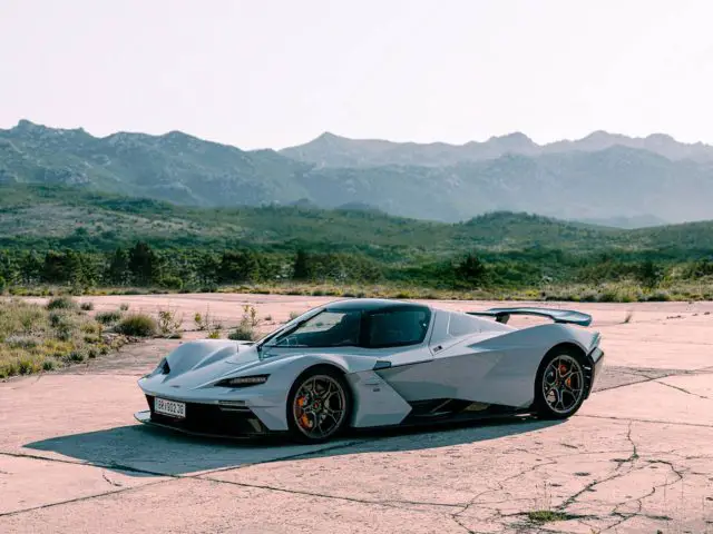 Een strakke zilveren sportwagen De KTM X-BOW staat geparkeerd op een gebarsten betonnen ondergrond met een bergachtig landschap op de achtergrond onder een heldere hemel.