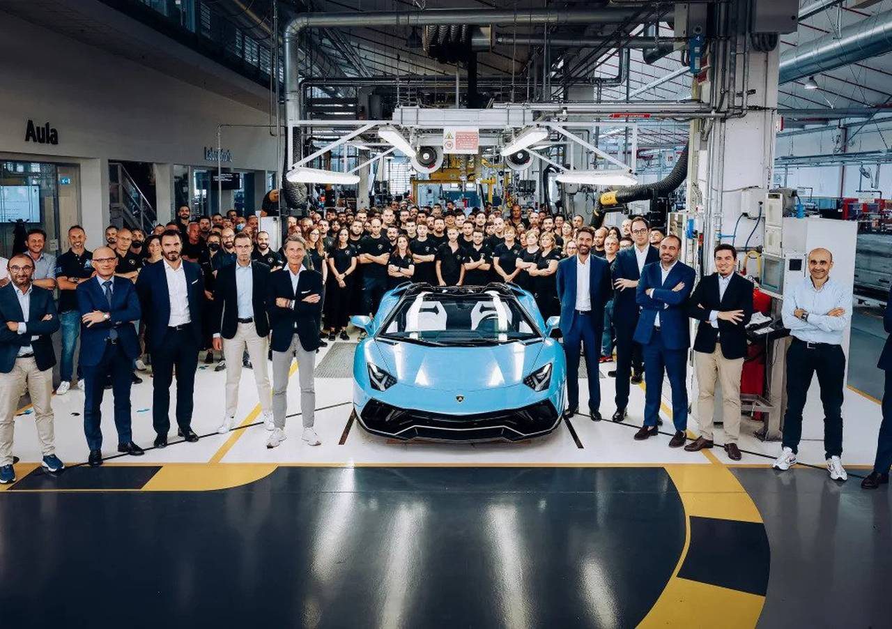Een groep mensen poseert rond een Aventador in een goed verlichte autofabriek. De blauwe sportwagen staat centraal onder plafondverlichting, met op de achtergrond diverse apparatuur en werkplekken zichtbaar.