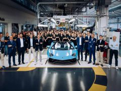 Een groep mensen poseert rond een Aventador in een goed verlichte autofabriek. De blauwe sportwagen staat centraal onder plafondverlichting, met op de achtergrond diverse apparatuur en werkplekken zichtbaar.