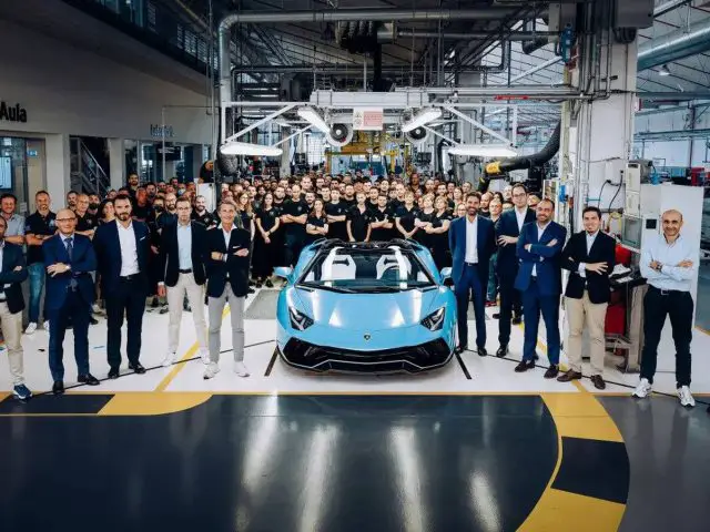 Een groep mensen poseert rond een Aventador in een goed verlichte autofabriek. De blauwe sportwagen staat centraal onder plafondverlichting, met op de achtergrond diverse apparatuur en werkplekken zichtbaar.