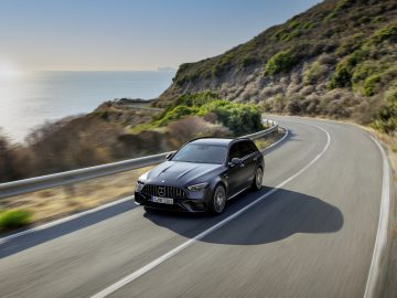 Een strakke Mercedes-AMG C 63 S E Performance rijdt over een bochtige kustweg met links de zon ondergaand boven de oceaan en rechts groene heuvels.