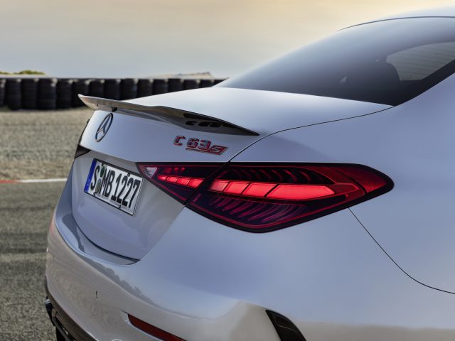 Het achteraanzicht van een witte luxeauto wordt getoond met de kenmerkende achterlichten en het modellabel "Mercedes-AMG C 63 S E Performance" zichtbaar nabij de achterband van het voertuig. De achtergrond omvat een baan en bandenbarrières.