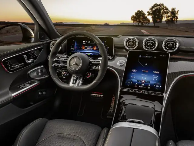 Interieur van een moderne Mercedes-AMG C 63 S E Performance met een digitaal dashboard, multifunctioneel stuur en centraal touchscreen-display met verschillende bedieningselementen. De scène speelt zich af tijdens zonsondergang, met buiten een racecircuit zichtbaar.