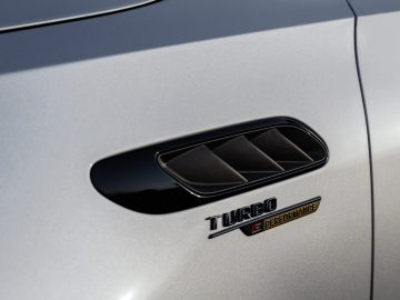 Close-up van een Mercedes-AMG C 63 S E Performance ventilatierooster aan de zijkant met de woorden 'Turbo E Performance' in zwarte en rode tekst op een strakke metallic zilveren lak.