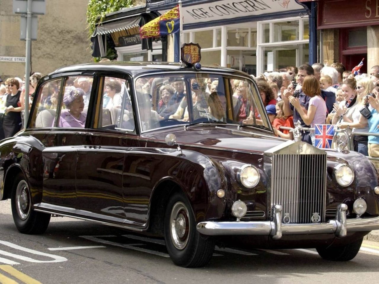 Een vintage Rolls-Royce limousine met passagiers rijdt door een drukke straat met toeschouwers en vlaggen op de achtergrond, wat doet denken aan een grote optocht voor Koning Charles.