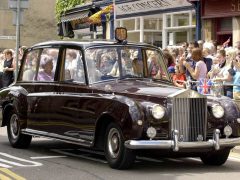Een vintage Rolls-Royce limousine met passagiers rijdt door een drukke straat met toeschouwers en vlaggen op de achtergrond, wat doet denken aan een grote optocht voor Koning Charles.