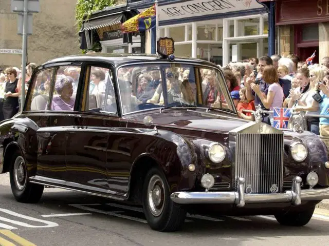 Een vintage Rolls-Royce limousine met passagiers rijdt door een drukke straat met toeschouwers en vlaggen op de achtergrond, wat doet denken aan een grote optocht voor Koning Charles.