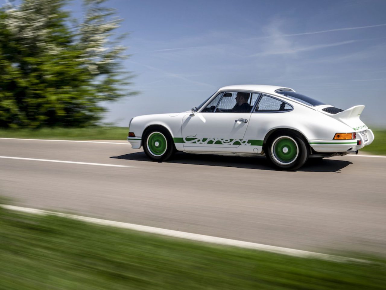 Een witte klassieke Porsche 911 Carrera 2.7 RS met groene accenten rijdend op een landelijke weg, omgeven door weelderig groen en een helderblauwe lucht op de achtergrond.
