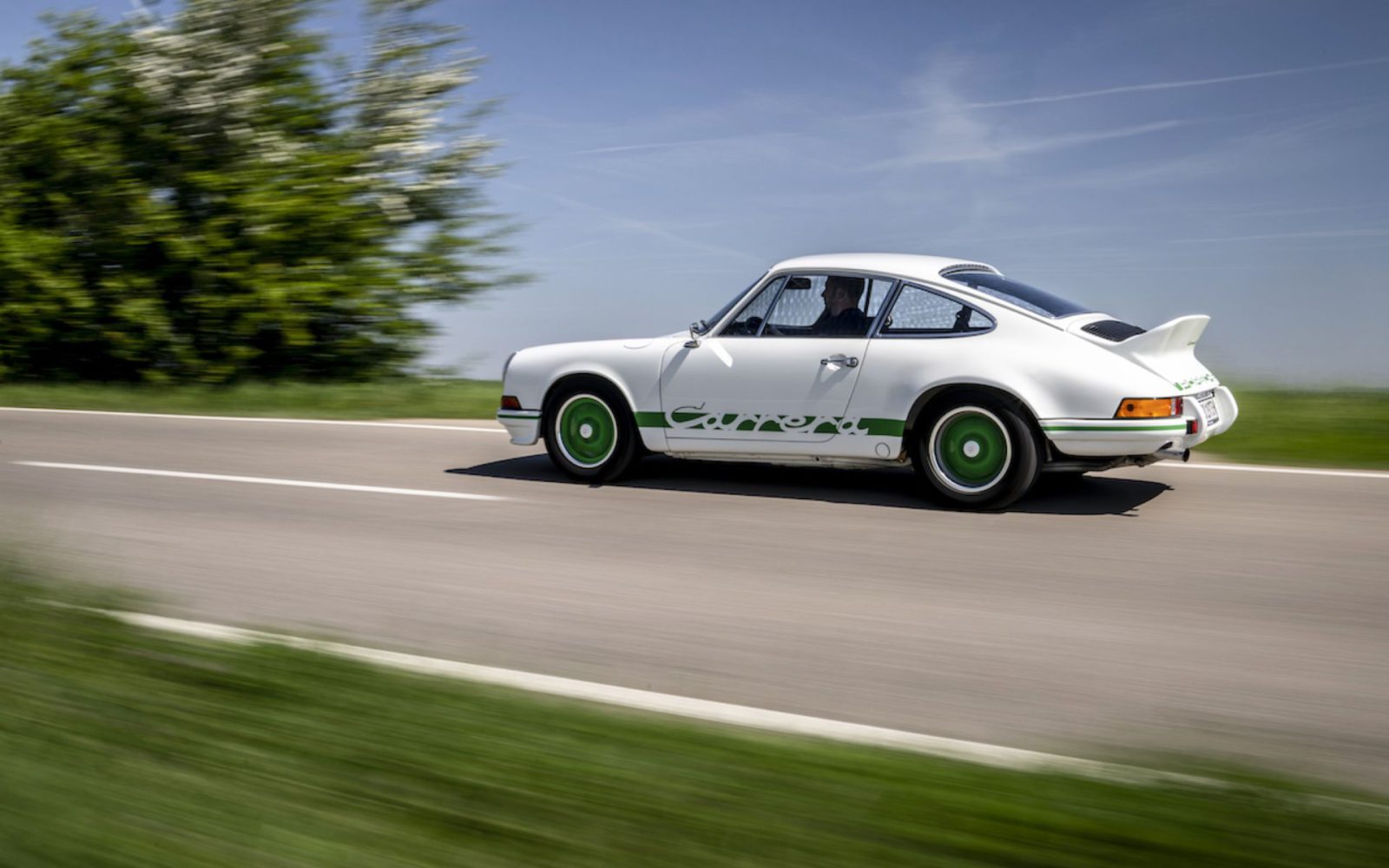 Een witte klassieke Porsche 911 Carrera 2.7 RS met groene accenten rijdend op een landelijke weg, omgeven door weelderig groen en een helderblauwe lucht op de achtergrond.