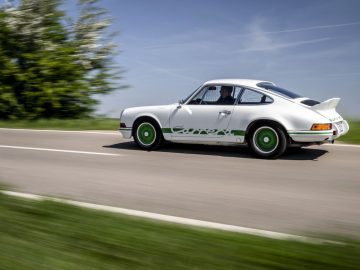 Een witte klassieke Porsche 911 Carrera 2.7 RS met groene accenten rijdend op een landelijke weg, omgeven door weelderig groen en een helderblauwe lucht op de achtergrond.