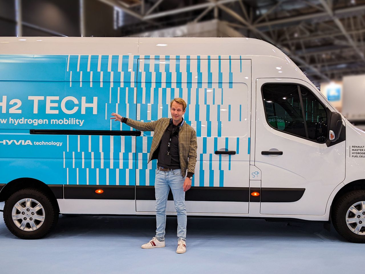 Een persoon staat naast een wit busje met de logo's "H2 TECH new waterstofmobiliteit" en "HYVIA technology" op de zijkant, waarmee het innovatieve gebruik van waterstof wordt getoond.