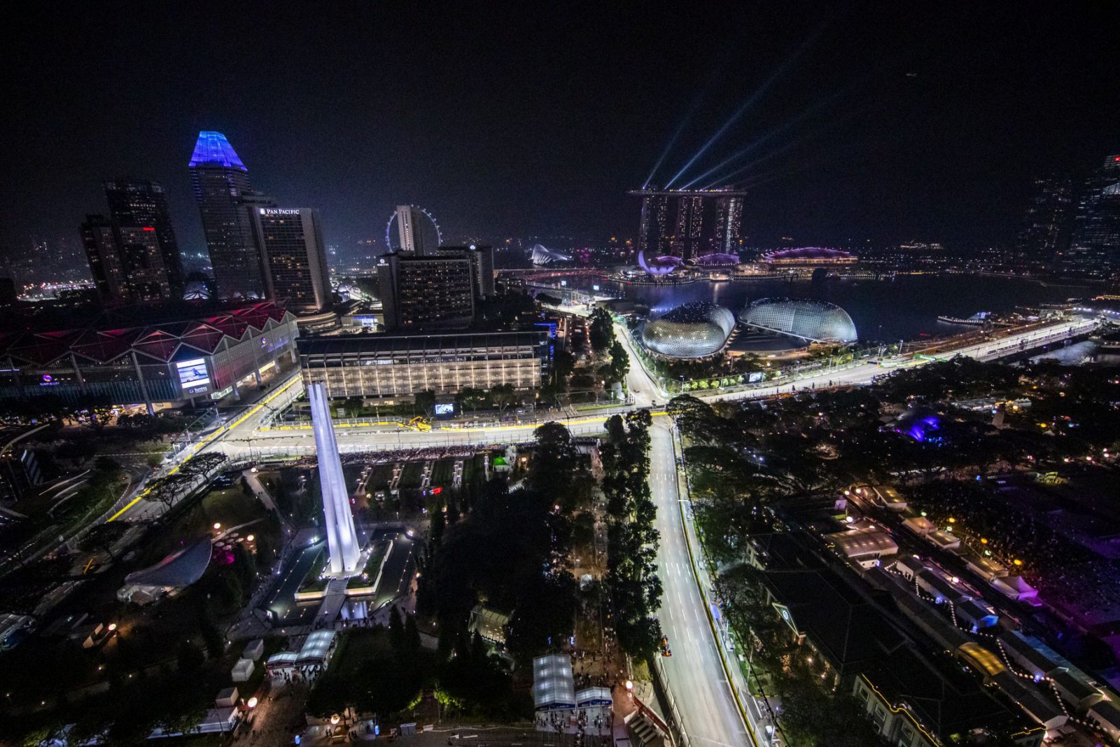Nachtelijk luchtfoto van een goed verlichte stad met wolkenkrabbers, bouwwerken aan het water, een reuzenrad en een netwerk van verlichte wegen en bruggen dat doet denken aan de oogverblindende Tijden van de GP Singapore 2022.