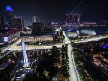 Nachtelijk luchtfoto van een goed verlichte stad met wolkenkrabbers, bouwwerken aan het water, een reuzenrad en een netwerk van verlichte wegen en bruggen dat doet denken aan de oogverblindende Tijden van de GP Singapore 2022.