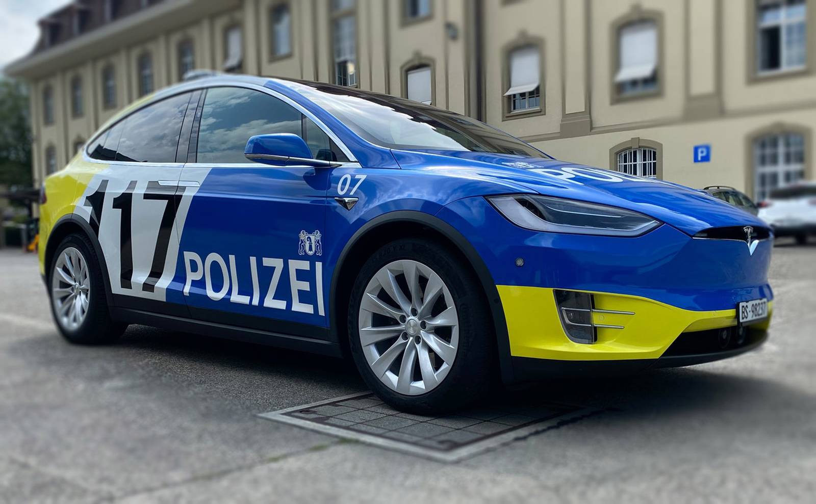 Een strakke Tesla Model X-politieauto staat geparkeerd buiten een gebouw, geschilderd in blauw en geel met "POLIZEI" en het nummer 17 duidelijk aangegeven op de zijkant.