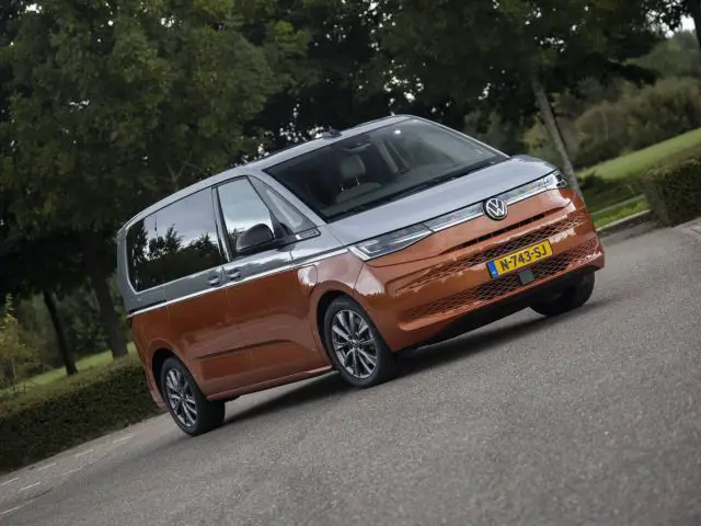 Een tweekleurige oranje en grijze Volkswagen Multivan geparkeerd op een verharde weg met groen op de achtergrond.