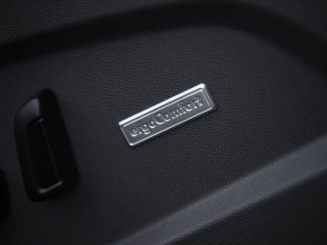 Close-up van een Volkswagen Multivan-autostoeltje met een zilveren "ergoComfort"-badge met aangrenzende bedieningsknoppen op een gestructureerd oppervlak.