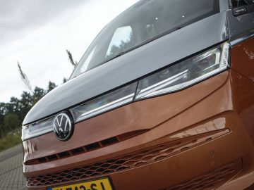 Close-up van de voorkant van een tweekleurige Volkswagen Multivan met een opvallend logo op de grille, buiten geparkeerd. Het bovenste gedeelte is zilver en het onderste gedeelte is oranje. De koplampen en de kentekenplaat aan de voorkant zijn zichtbaar.