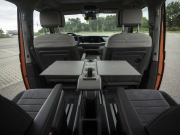 Binnenaanzicht van een Volkswagen Multivan met twee voorstoelen, een opvouwbare middentafel en twee kapiteinsstoelen achterin, allemaal bekleed met grijze stof, met een open parkeerplaats en bomen zichtbaar door de ramen.