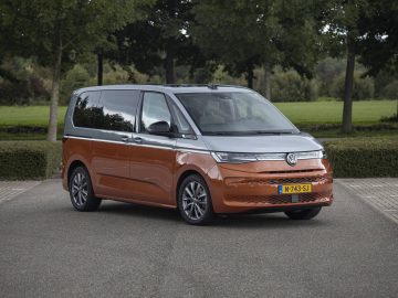 Een tweekleurige oranje en zilveren Volkswagen Multivan staat geparkeerd op een verhard terrein, met bomen en groen op de achtergrond.