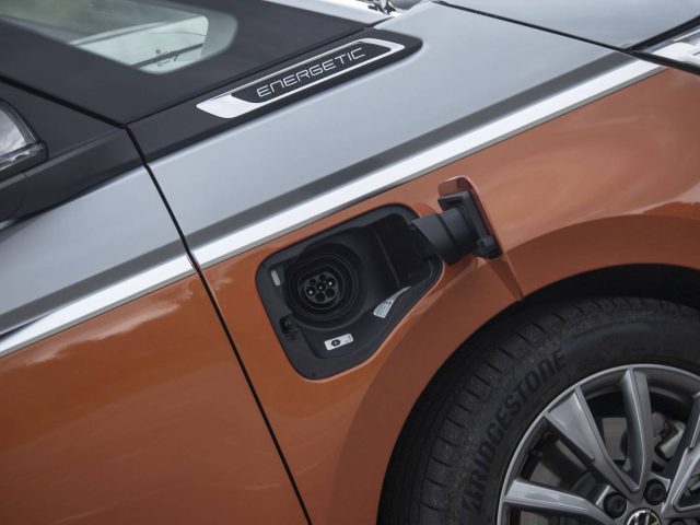 Close-up van de oplaadpoort van een elektrische auto aan de linkerkant, boven het achterwiel, met een open klep en het label "Energetic" zichtbaar op de carrosserie van de Volkswagen Multivan.