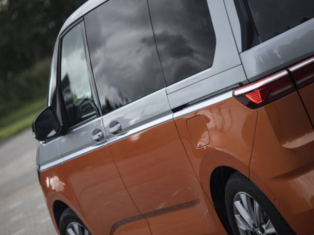 Zijaanzicht van een Volkswagen Multivan in oranje en grijs, met de nadruk op de achterruit en een rood achterlicht tegen een onscherpe buitenachtergrond.