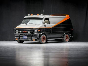 Een zwarte GMC-bestelwagen met oranje details en aangepaste wielen, die doen denken aan de iconische rit van het A-Team, staat binnen geparkeerd tegen een donkere achtergrond.