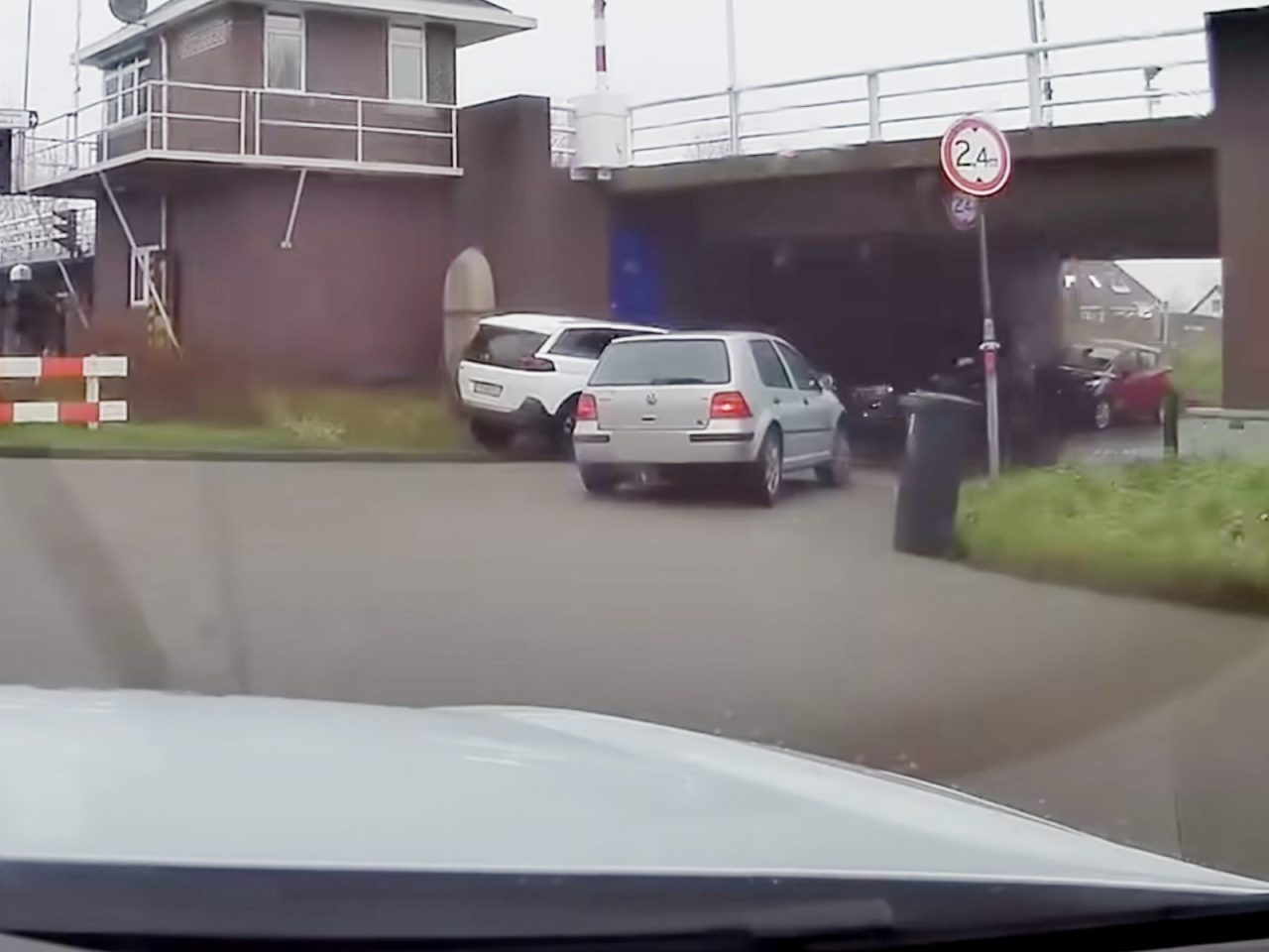 Verschillende auto's, waaronder een Politie Volkswagen Golf, staan geparkeerd bij een bakstenen gebouw en onder een brug, met een witte SUV onhandig schuin op een verhoogd grasveld. Bij de brug is een bord met een hoogtebeperking van 2,4 meter zichtbaar.