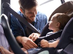 Een man zet een baby vast in een autostoeltje terwijl hij lacht naar het blije gezicht van het kind. De baby draagt een gestreept shirt.