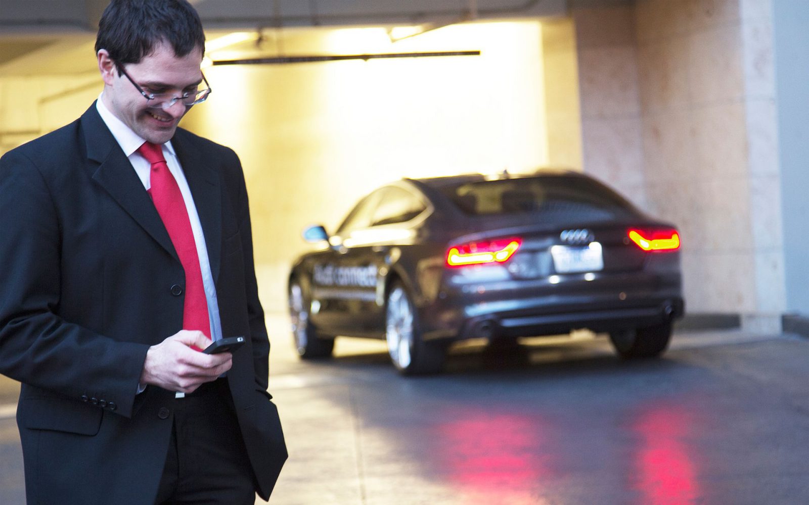 Een man in pak en rode das staart naar zijn telefoon, terwijl hij naast een geparkeerde grijze Audi staat, en controleert misschien zijn boetestatistieken.