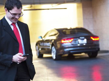 Een man in pak en rode das staart naar zijn telefoon, terwijl hij naast een geparkeerde grijze Audi staat, en controleert misschien zijn boetestatistieken.