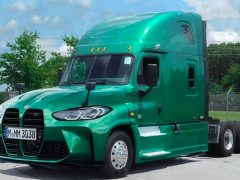 Een groene semi-vrachtwagen met een uniek grille-ontwerp, dat doet denken aan BMW-vrachtwagens, geparkeerd op een buitenterrein met bomen en een hek op de achtergrond.