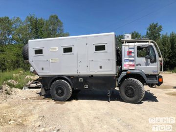 Op een onverharde weg staat een stoere, zilverkleurige expeditietruck geparkeerd, ideaal voor wie een camper wil huren. Het voertuig is voorzien van verhoogde ramen, offroad-banden en een boxy cabine. Op de achtergrond zijn bomen en groen zichtbaar.