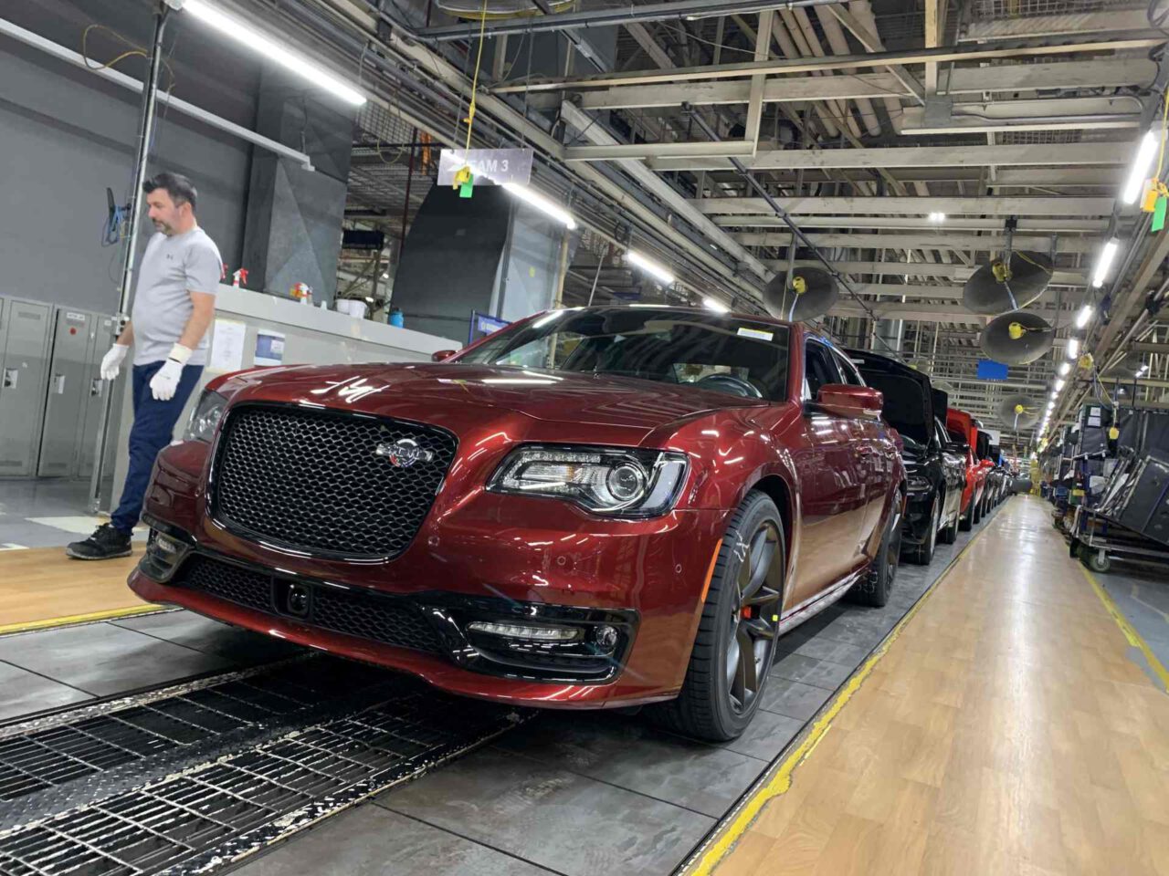 Een rij rode Chrysler 300-auto's in verschillende productiefasen beweegt zich door een lopende band in een fabriek, met vlakbij een arbeider in een wit overhemd en handschoenen.