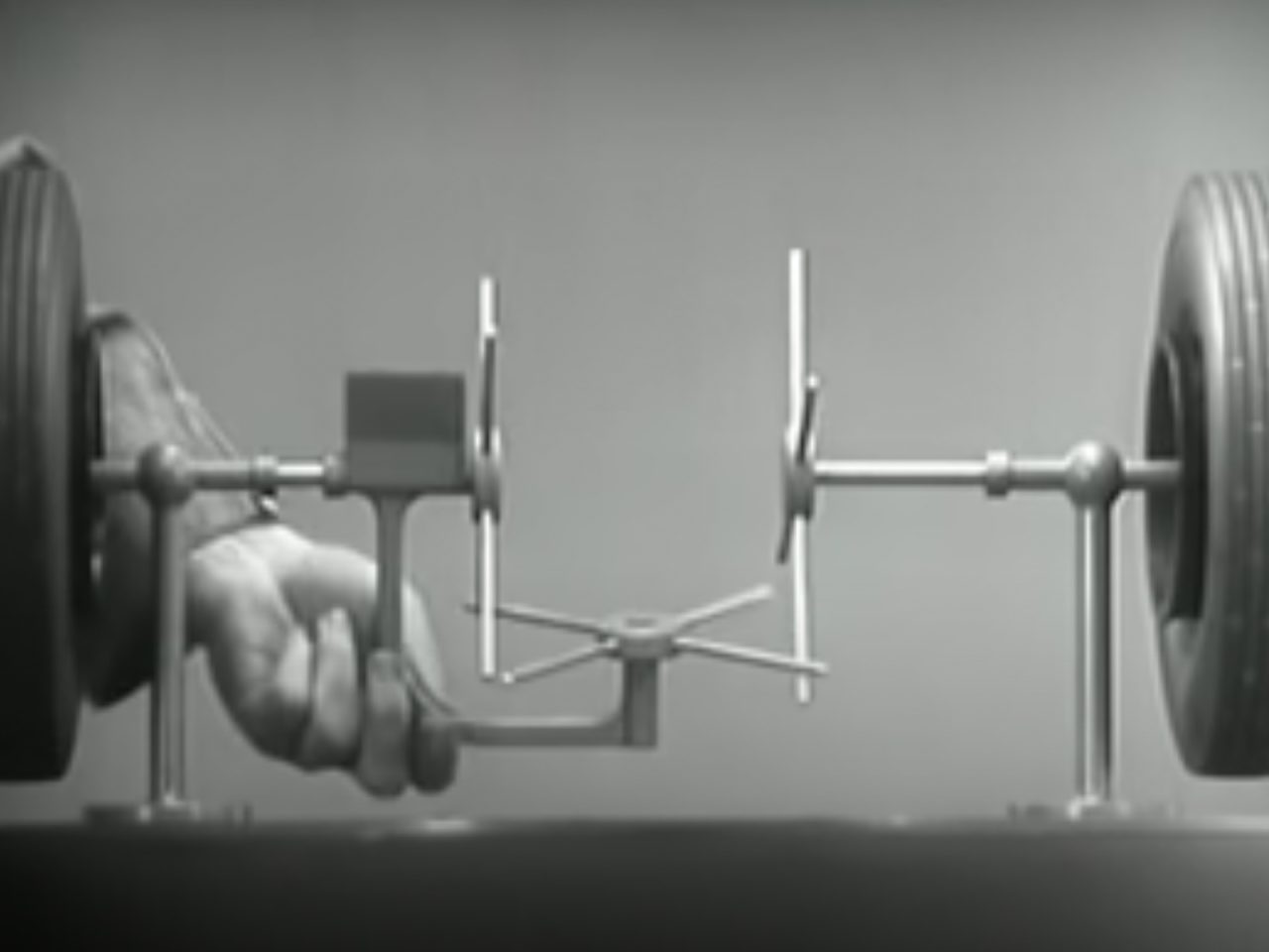 Een zwart-wit afbeelding van de hand van een persoon die de as van een model vasthoudt en de werking van het differentieel versnellingssysteem van een voertuig demonstreert, met aan weerszijden twee wielen gemonteerd.