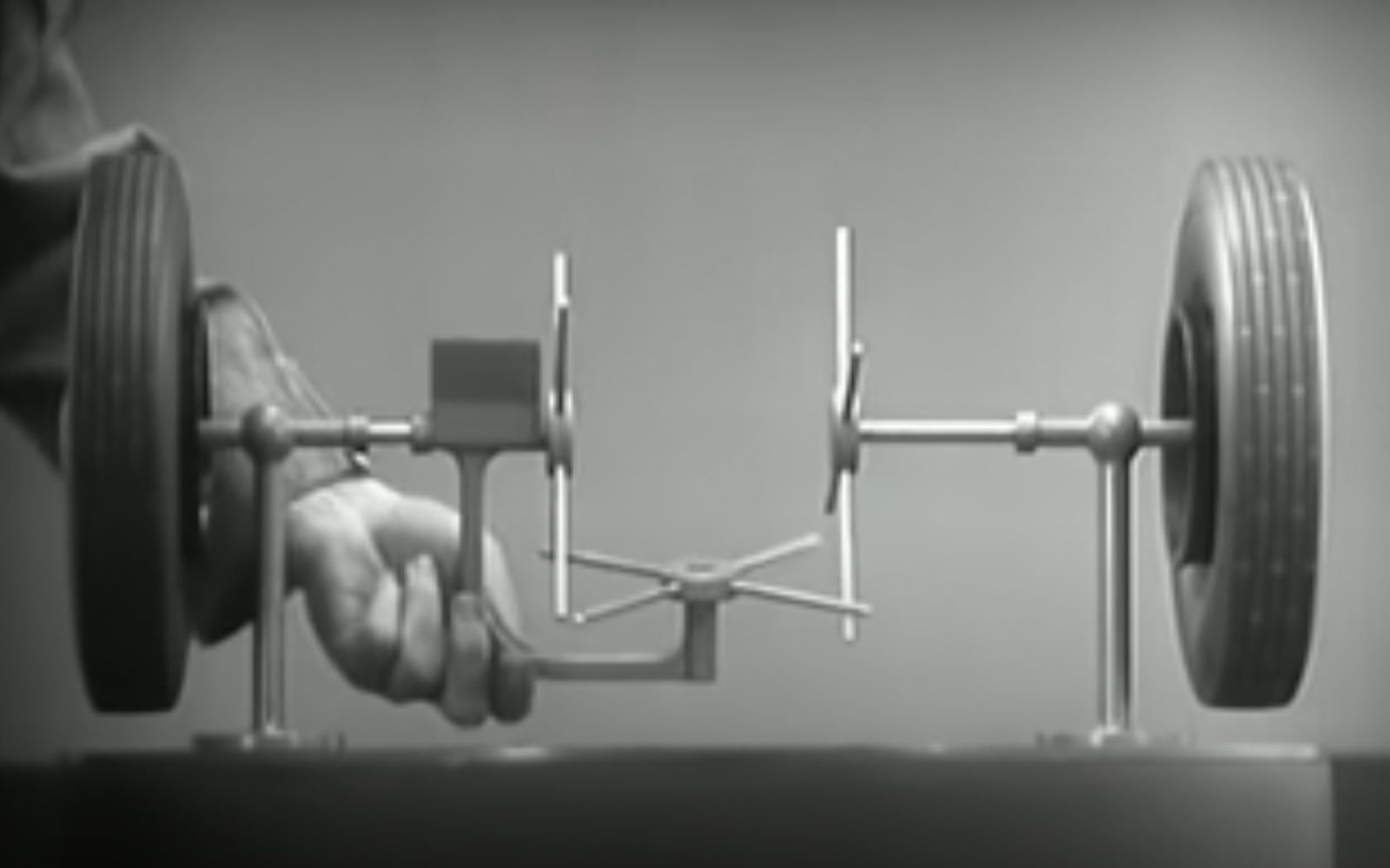 Een zwart-wit afbeelding van de hand van een persoon die de as van een model vasthoudt en de werking van het differentieel versnellingssysteem van een voertuig demonstreert, met aan weerszijden twee wielen gemonteerd.