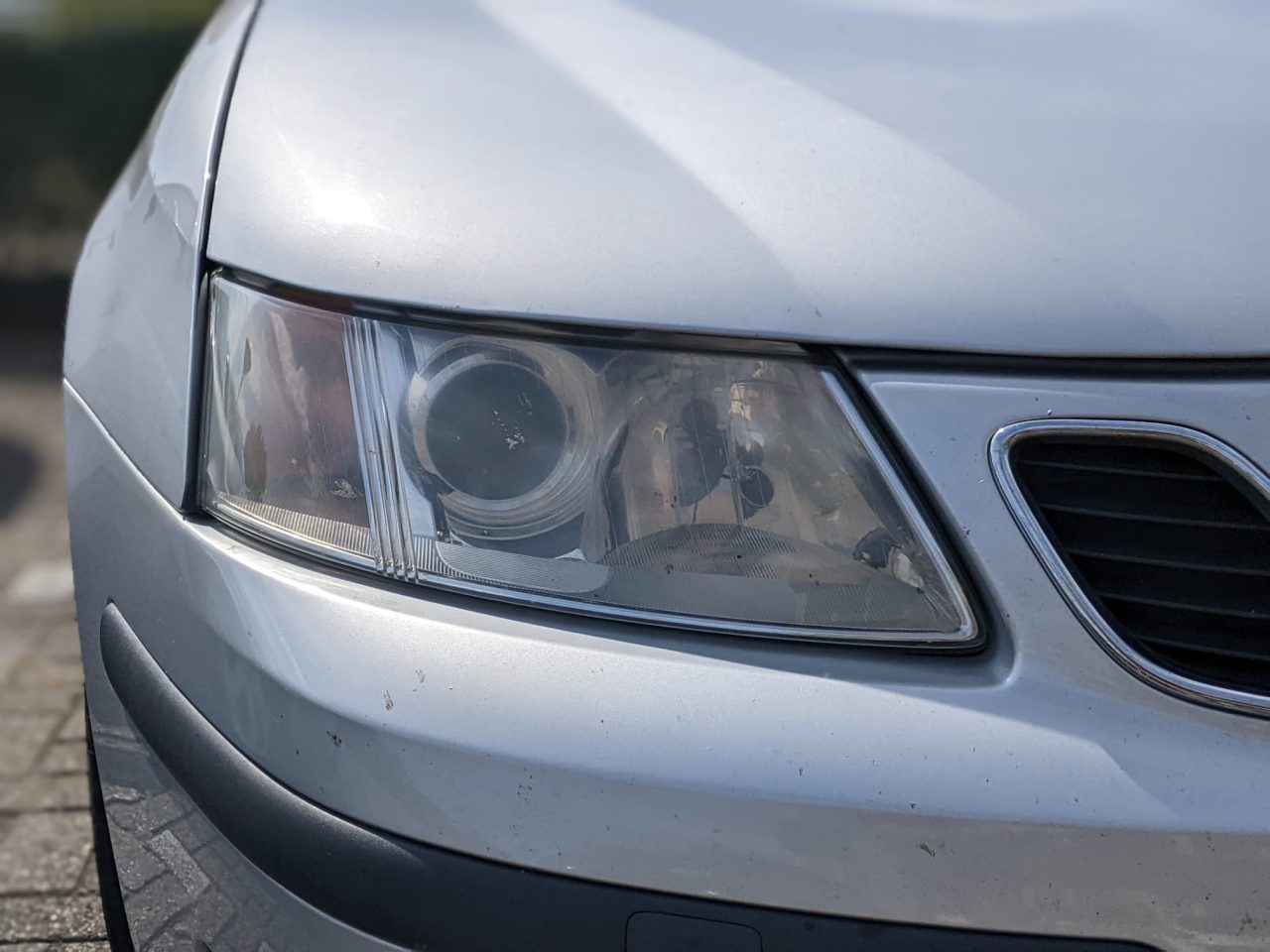 Close-up van de koplamp linksvoor van een zilveren auto en een deel van de bumper, met wat klein vuil en slijtage aan de doffe koplampen.