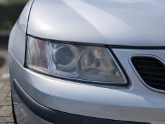 Close-up van de koplamp linksvoor van een zilveren auto en een deel van de bumper, met wat klein vuil en slijtage aan de doffe koplampen.