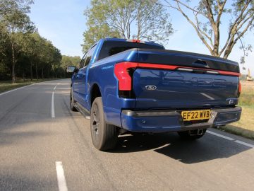 Een blauwe Ford F-150 Lightning pick-up rijdt op een tweebaansweg, omgeven door groen en bomen. Op het kenteken staat "EF 22 NVB.