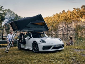 Op een mooie buitenlocatie staat een witte Porsche, voorzien van een Porsche Tequipment daktent. Er zijn twee mensen in de buurt: een staat op een ladder bij de tent en een ander zit op de grond bij een serene vijver.