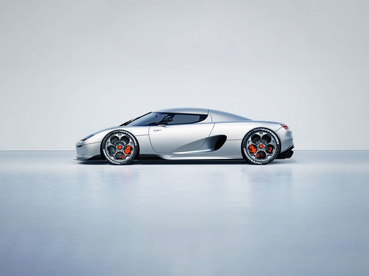 Een strakke, moderne zilveren Koenigsegg-sportwagen met een aerodynamisch ontwerp en zwarte wielen geaccentueerd met oranje accenten, weergegeven tegen een effen, lichtgekleurde achtergrond.