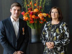 Een man in pak met een medaille staat naast een vrouw in een jasje met patronen voor een hoge vaas met kleurrijke bloemen, wat doet denken aan de elegantie die je zou associëren met de voorname aanwezigheid van Max Verstappen.