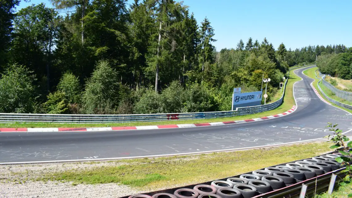 Een racecircuit met een scherpe bocht omzoomd door rode en witte stoepranden, omgeven door bomen en groen onder een heldere hemel. Een deel van de baan bevat teksten en tekeningen. Er zijn zwarte banden langs de rand, wat ondanks de hoge kosten bijdraagt aan het authentieke Nürburgring-gevoel.