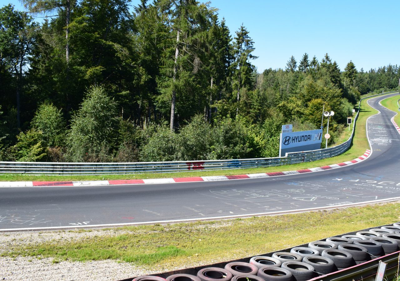 Een racecircuit met een scherpe bocht omzoomd door rode en witte stoepranden, omgeven door bomen en groen onder een heldere hemel. Een deel van de baan bevat teksten en tekeningen. Er zijn zwarte banden langs de rand, wat ondanks de hoge kosten bijdraagt aan het authentieke Nürburgring-gevoel.