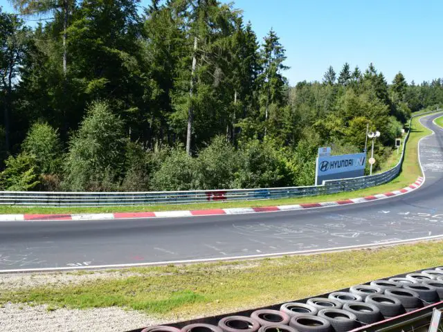 Een racecircuit met een scherpe bocht omzoomd door rode en witte stoepranden, omgeven door bomen en groen onder een heldere hemel. Een deel van de baan bevat teksten en tekeningen. Er zijn zwarte banden langs de rand, wat ondanks de hoge kosten bijdraagt aan het authentieke Nürburgring-gevoel.