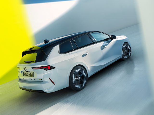 Een witte moderne Opel Astra GSe hatchback met een zwart dak is van achteren te zien, rijdend op een heldere weg met gele en witte muren op de achtergrond.