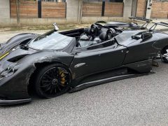 Aan de kant van de weg staat een beschadigde zwarte Pagani Zonda-sportwagen met aanzienlijke schade aan de voor- en achterwielen. De auto heeft ernstige krassen en er ontbreken onderdelen. Op de achtergrond zijn barrières en een hek zichtbaar.