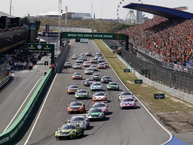 Een groep raceauto's, waaronder een strakke Porsche, concurreert op een circuit omringd door toeschouwers op tribunes tijdens een autosportevenement op een heldere dag.