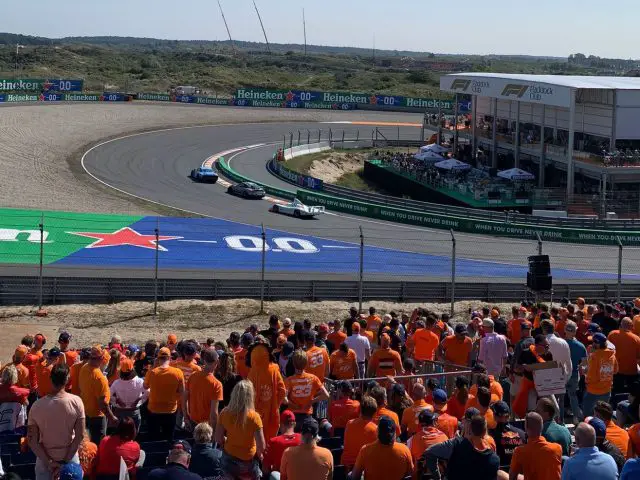 Een menigte toeschouwers in oranje shirts kijkt naar een Formule 1-race op een circuit, waarbij drie auto's, waaronder een Porsche, een bocht nemen nabij een gebied met het merk Heineken.