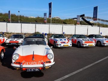 Buiten staat een rij vintage politieauto's geparkeerd. Op de voorgrond staat een wit-oranje Porsche met "RIJKSPOLITIE" op de voorkant.