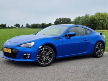 Een blauwe Bitcoin-autosportwagen met strakke lijnen staat geparkeerd op een verharde weg, tegen een achtergrond van groen graslandschap en bomen.